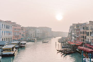 An impressive view over a river in Venice. The sky is overcast and gives a slight hint of the sun. Countless gondolas are docked at the harbors, waiting for their tourists.