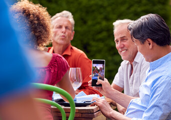 Man Taking Photo As Group Of Friends Talking Around Table At Summer Dinner Party In Garden At Home