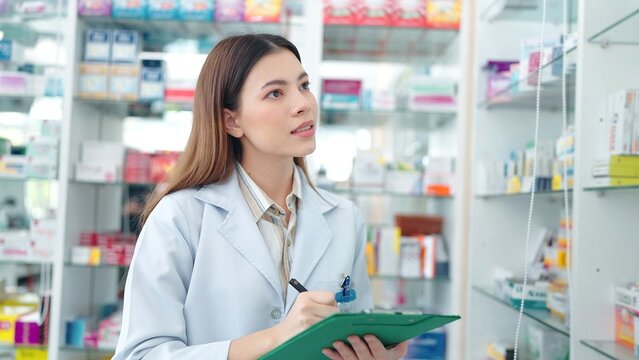 Professional Asian woman pharmacist rechecks products in pharmacy and using noteboard to write information