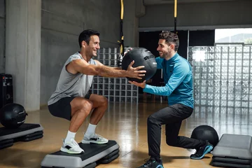 Tuinposter Fitness personal trainer helping man with squatting pose with medicine ball © BASILICOSTUDIO STOCK
