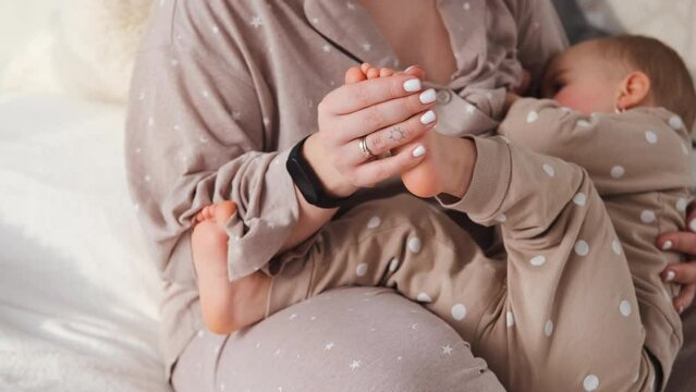 Mother caresses her baby's legs during breastfeeds, close up of mom massages baby child's feet while breastfeeding her