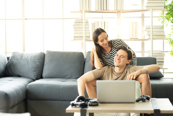 A couple sitting together in the living room enjoy relaxing at home. Young man sits on floor while his girlfriend sits on couch watching online movie from laptop computer. Couple leisure activity
