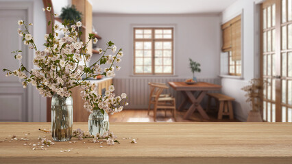 Wooden table, desk or shelf close up with branches of cherry blossoms in glass vase over blurred view of farmhouse wooden kitchen, minimalist interior design concept
