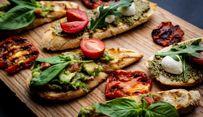 Set of bruschettas with pesto, guacamole, grilled tomatoes, mozzarella cheese, basil and arugula on wooden board. Italian tosted bread with vegetables and traditional sauces