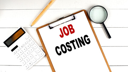 JOB COSTING words on clipboard, with calculator, magnifier and pencil on the white wooden background