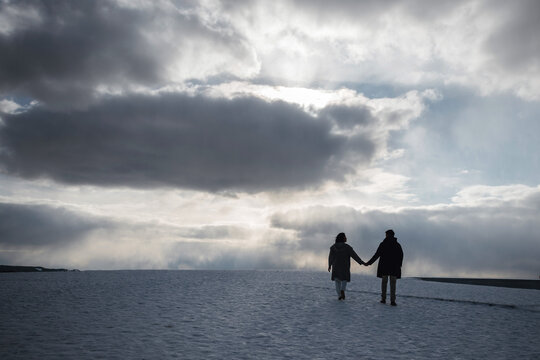 Rear view of young couple in winter clothes walking holding hands on the snow surface on the rooftop.