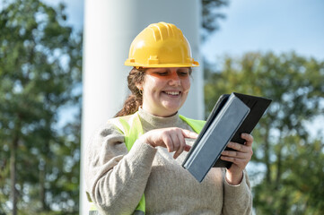 Close-up female engineer with brown curly hair is smiling while typing on a digital tablet in front of a wind turbine during daylight