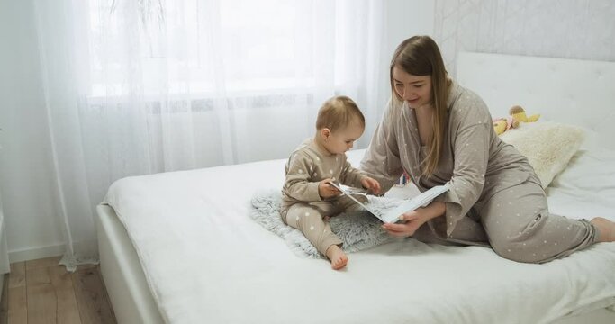 Baby child with her young mother reading a book on the bed in the bedroom