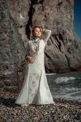Fototapeta premium Middle aged woman looks good with blond hair, boho style in white long dress on the beach decorations on her neck and arms.