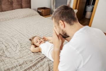 A father is playing peek a boo with his baby girl while she lying on the bed and laughing.