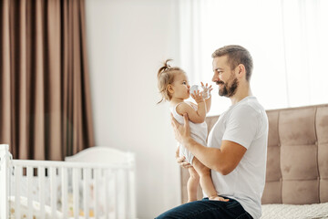 A father is holding his daughter in hands while she is looking at him and drinking water from bottle