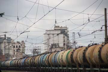a row of train cars sitting on a train track.a huge number of wagons with oil products, wires in the sky, an old factory in the background. Transport and energy, fossil fuels