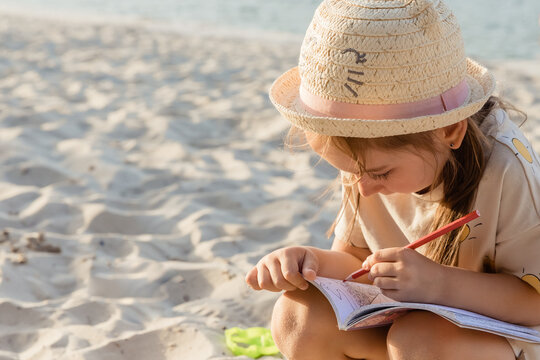 A little cute girl sits on the beach on vacation and draws on paper with colored pencils. Family on a summer weekends. Little girl drawing. Happy summer vacation concept