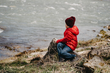 A little boy in a red jacket and a hat sits on a log by the river.