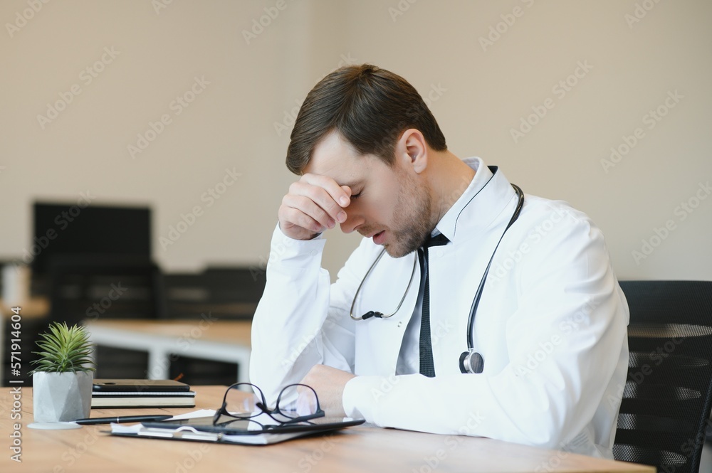 Wall mural stressed male doctor sat at his desk. mid adult male doctor working long hours. overworked doctor in