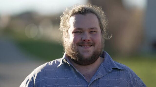 One happy overweight young man smiling at camera standing outdoors in sunlight. Natural casual male caucasian person looking at camera