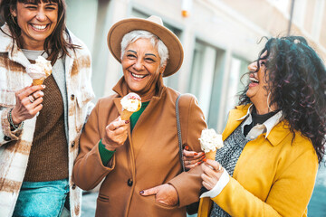 Three mature women eating ice cream cone outside - Older female friends having fun walking on city...