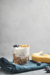 Granola cereal oatmeal with white yogurt, blueberries and banana fruits in a glass on a blue napkin, grey background