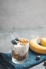 Granola cereal oatmeal with white yogurt, blueberries and banana fruits in a glass on a blue napkin, grey background