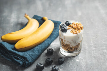 Granola cereal oatmeal with white yogurt, blueberries and banana fruits in a glass on a blue napkin, grey background