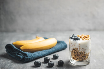 Granola cereal oatmeal with white yogurt, blueberries and banana fruits in a glass on a blue napkin, grey background