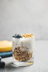 Granola cereal oatmeal with white yogurt, blueberries and banana fruits in a glass on a grey background