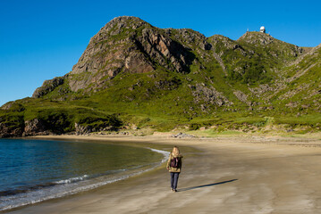 Young woman with a backpack walks along an empty sandy ocean beach. Town and mountains. Beautiful nature landscape. Lonely girl. Wanderlust. Travel, adventure, lifestyle. Explore North, Norway