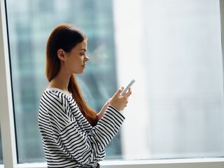 Obraz premium Woman holding her phone and looking and smiling at it near the window of the house, online shopping from home