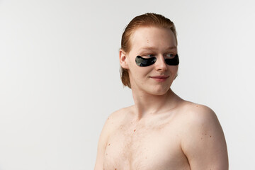 Taking care after skin with cosmetics. Young redhead man with eye patches posing shirtless against grey studio background. Concept of men's health, body and skin care, hygiene and male cosmetology