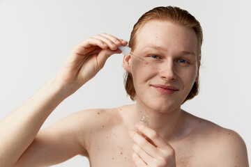 Young man taking care after skin. Redhead model posing against grey studio background. Applying face serum for hydration. Concept of men's health, body and skin care, hygiene and male cosmetology