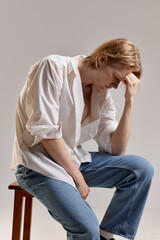 Portrait of young redhead man posing in white shirt and jeans sitting on chair with thoughtful face on grey studio background. Concept of men's health, body and skin care, hygiene and male cosmetology