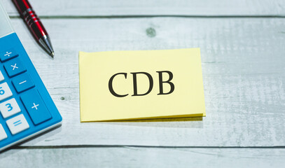 A sigla CDB de Certificado de Depósito Bancário escrita em um pedaço de papel que está sobre uma mesa de madeira. Uma calculadora e uma caneta na composição. Economia brasileira e investimentos. - obrazy, fototapety, plakaty