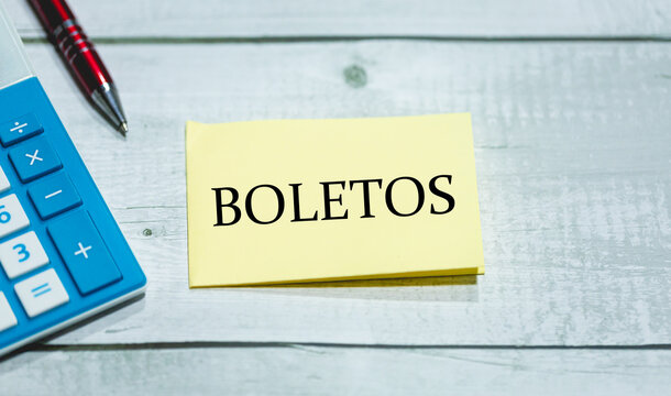 A palavra boletos em Portugu&ecirc;s do Brasil escrita em um peda&ccedil;o de papel que est&aacute; sobre uma mesa de madeira. Uma calculadora e uma caneta na composi&ccedil;&atilde;o. Economia brasileira e finan&ccedil;as.