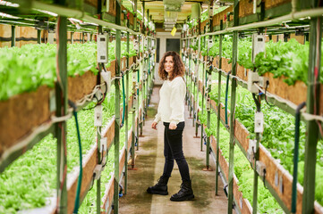 Portrait of female person looking at green leafy plants in greenhouse. Full length of joyful young woman reaching out hand to leafy greens and smiling, standing in aisle between shelves with plants. - Powered by Adobe