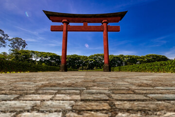 Torii, gate of Japanese shrine. 3D illustration of a wooden Japanese torii gate painted in red and black colors on a stone path with bushes on the side and trees in the background on a sunny day