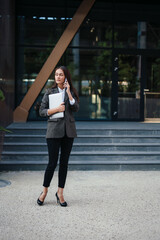 Young brunette woman holding laptop, talking on mobile phone call while standing outdoors in city. Business woman or female freelancer, working outdoors