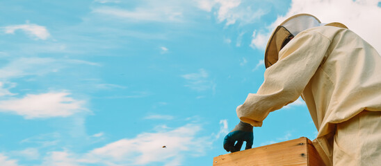 Banner. A beekeeper in a protective suit works with bees against the background of a blue sky. A...