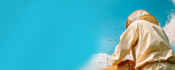 Banner. A beekeeper in a protective suit works with bees against the background of a blue sky. A...