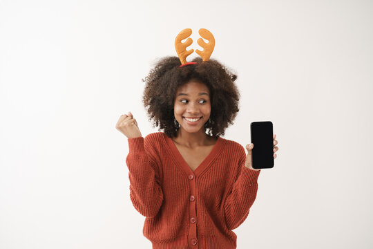 Smiling African American young woman holding smart phone up with happiness, happy yes gesture, exciting wearing reindeer hairband, festive season, holiday, Christmas shopping. Advertisement promotion