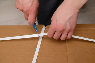 A man, a furniture assembler, opens a cardboard box with a clerical knife. The concept of unpacking the box.Close-up