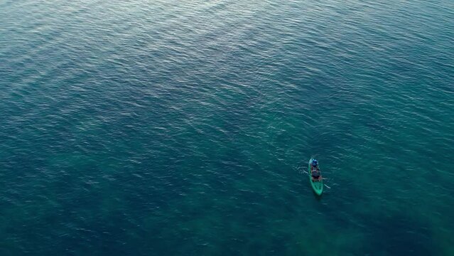 A drone view of a small narrow rowing boat that the two hikers on board are propelled by a two-bladed paddle. The kayak floats in turquoise sea at dawn, rising sun reflecting off sea surface of water.