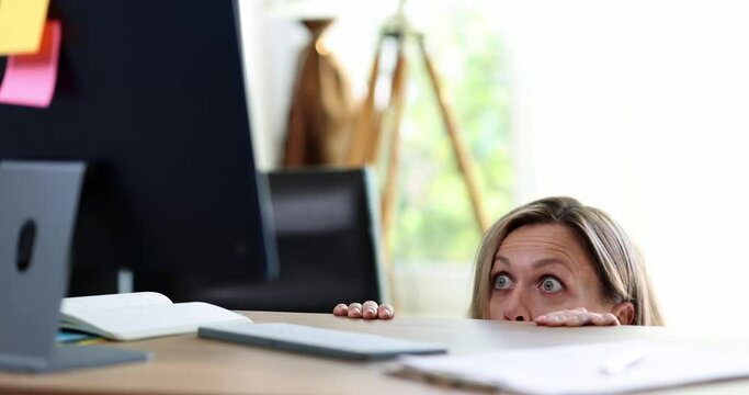 Portrait of surprised excited frightened woman peeking out from behind table and looking at computer screen