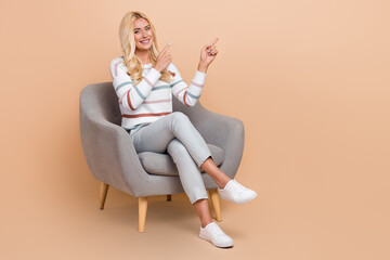 Fototapeta na wymiar Full length photo of attractive lady blonde hair wear striped sweater sit comfort armchair point fingers mockup isolated on beige color background