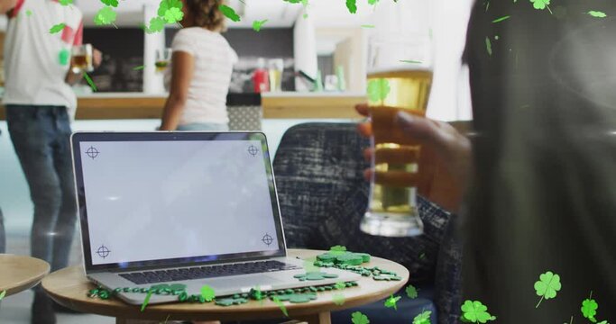 Animation of clover icons over diverse friends drinking beer and using laptop with copy space