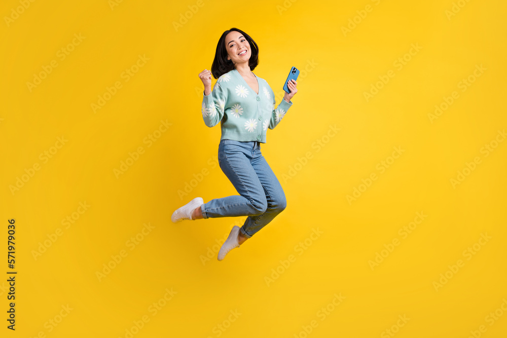 Sticker Full length photo of good mood nice girl dressed blue cardigan jeans jump up clench fist hold phone isolated on yellow color background