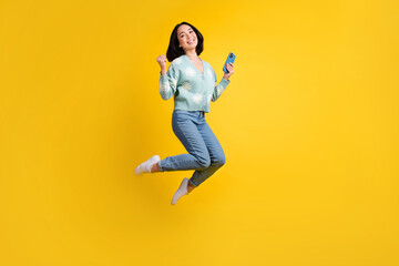 Fototapeta na wymiar Full length photo of good mood nice girl dressed blue cardigan jeans jump up clench fist hold phone isolated on yellow color background
