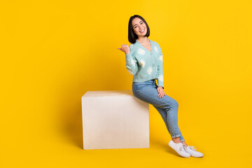 Full size photo of girlish satisfied woman dressed blue pullover directing empty space sit on cube isolated on yellow color background