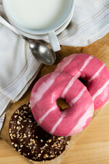 Top vertical view of donuts with chocolate and pink frosting over a wooden table, milk cup and white towe