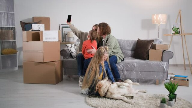 buying new apartment, joyful male and female parents together with cute son take selfie on smartphone camera while sitting on sofa, pretty girl plays with labrador while moving to cozy house