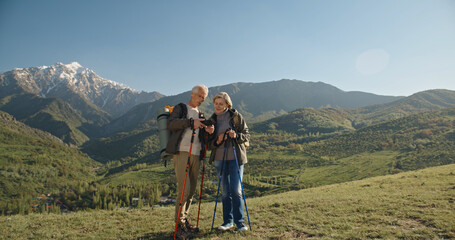 Fototapeta na wymiar Old couple having a hike in spring mountains, then stopping to take a picture on a smartphone. Senior caucasian family spending time together travelling after retirement - tourism concept 